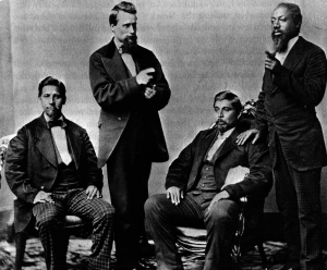 A photo of four men in suits, two of them sitting down in chairs.