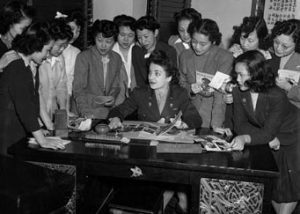 A group of women surround a table. They are Chinese American women who served the Army Air Force as "Air WACs." They performed aerial photo interpretation, air traffic control and weather forecasting.