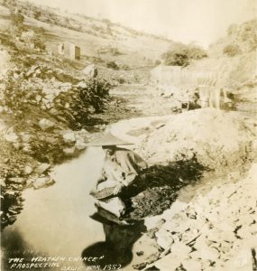 Chinese man in conical straw hat squatting on stream bank with gold pan in hand