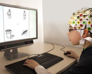A boy wearing an EEG cap uses a mouse and keyboard while looking at images on a computer monitor during an experiment.