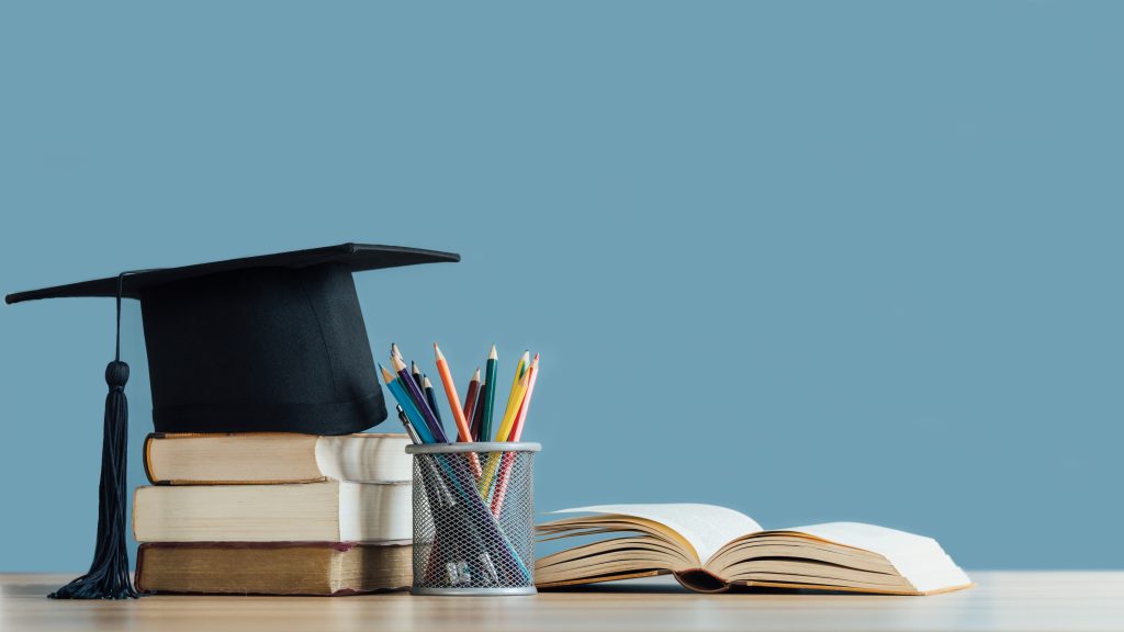 A mortarboard and graduation scroll on stack of books with pencils color in a pencil case on blue background.