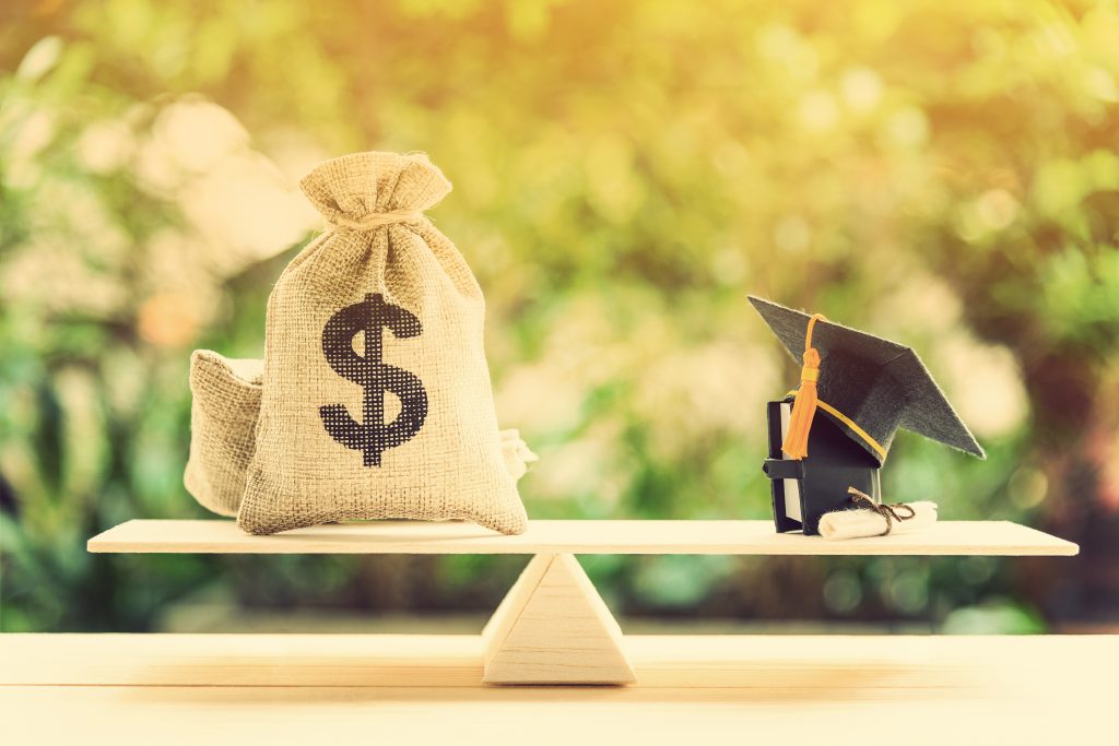 Cash in burlap bags and a black graduation cap with diploma and a book on opposite ends of a basic balance scale.