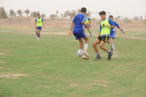 Six teenage boys play on a grass soccer field. Three wear yellow pinnies, two are in blue shirts, and the goal keeper stands in the goal in the background.