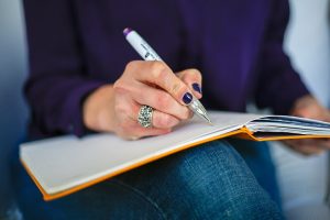 An image of the lower torso and upper crossed legs of a woman. She has a book spread across her lap and is writing in it with a pen.