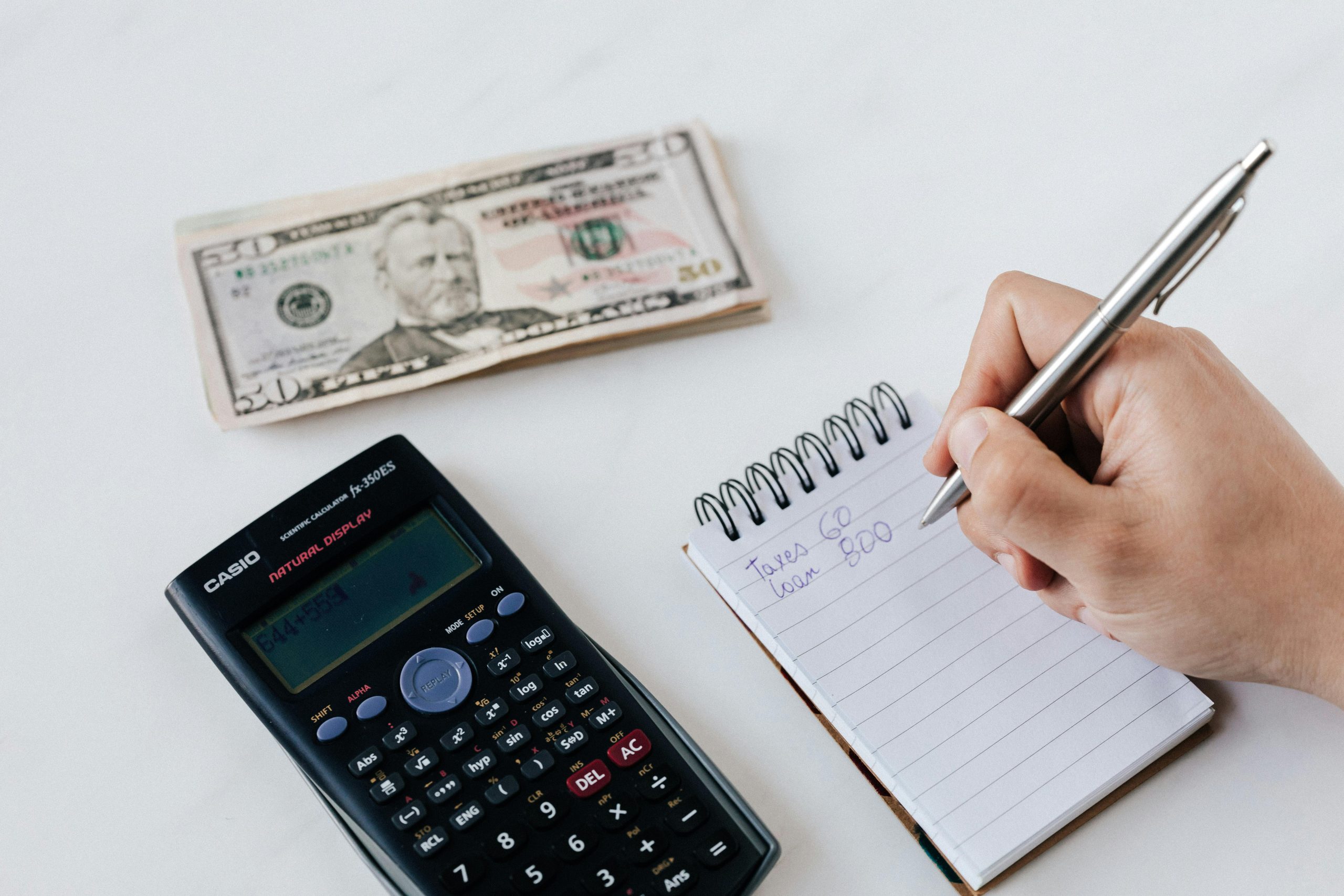 A photo of a calculator, stack of money and a hand writing notes on a notepad.