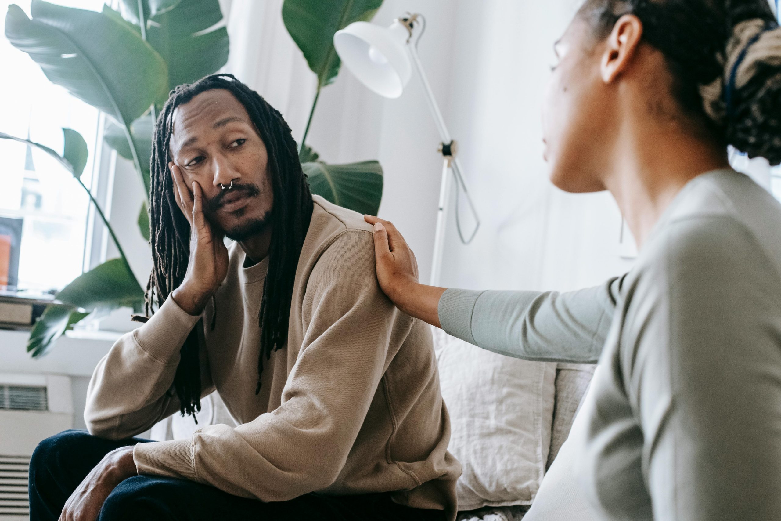 A woman consoles a man sitting next to her with placing a hand on his shoulder while he looks over to her with sadness in his face.