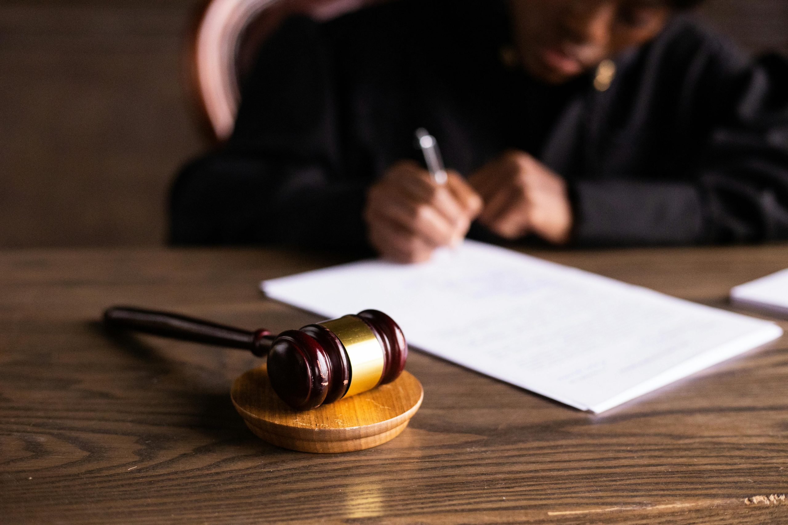 A gavel sits on a table with a judge in the background writing on a piece of paper.