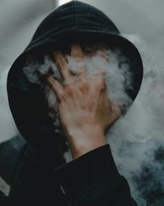 A man in a hoodie holding smoke over his face to obscure his identity.