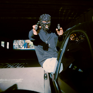 A man wearing a bandanna over his face sits out a car window pointing two guns at the camera