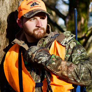 A man wearing camo and orange safety vests holds a rifle and leans against a tree