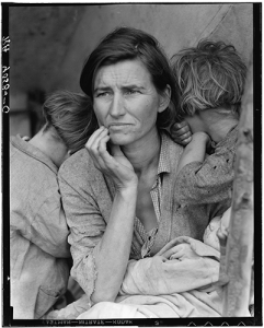 A careworn woman sits, surrounded by children clinging to her, looking into the distance.