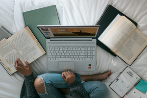 Overhead view of person using a laptop on a bed with several open books and notebooks lying around