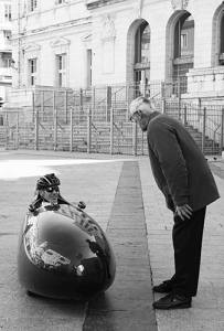 One man in a small, oval, really weird looking car, with another man standing over him and examining it.