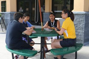 Four students with papers and books sit around an outdoor table.