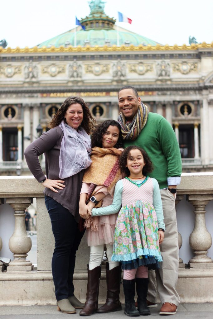 Photo of a family standing in front of a building. Mother, father, and two daughters are smiling.