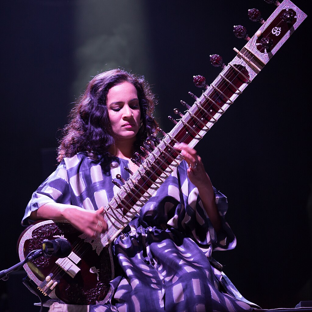 A woman playing a sitar on stage, illuminated by stage lighting.