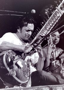 A black and white image of a musician playing a sitar on stage, with intricate designs on the instrument and another musician in the background.