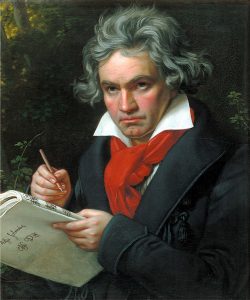 A man with gray hair and a red scarf holds a notebook and pencil, set against a dark, natural background.