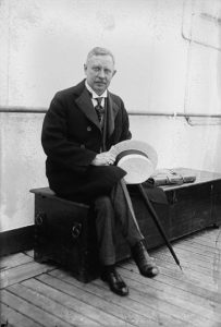 A man in early 20th-century attire sits on a wooden chest on a ship's deck, holding a straw boater hat.
