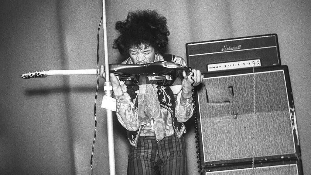 A person on stage holding a guitar upside down near their mouth, next to amplifiers.