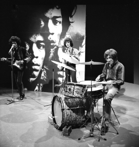 Black-and-white image of a band performing on stage with a large backdrop featuring a close-up face behind them.