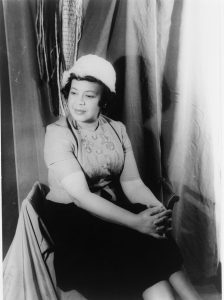 A seated woman in a hat and patterned blouse, with draped fabric and woven decorations in the background.