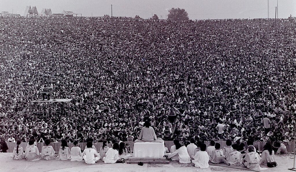 A black-and-white photo of a large crowd at an outdoor event, viewed from a stage with seated individuals facing the audience.