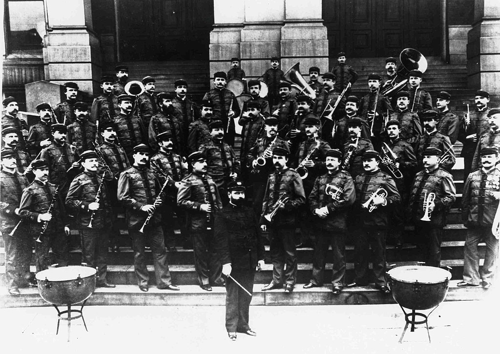 Historical military band in uniform standing on steps, with a conductor at the front and timpani drums to either side.