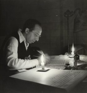 Person writing at a desk by candlelight, surrounded by sheets of paper and writing tools.