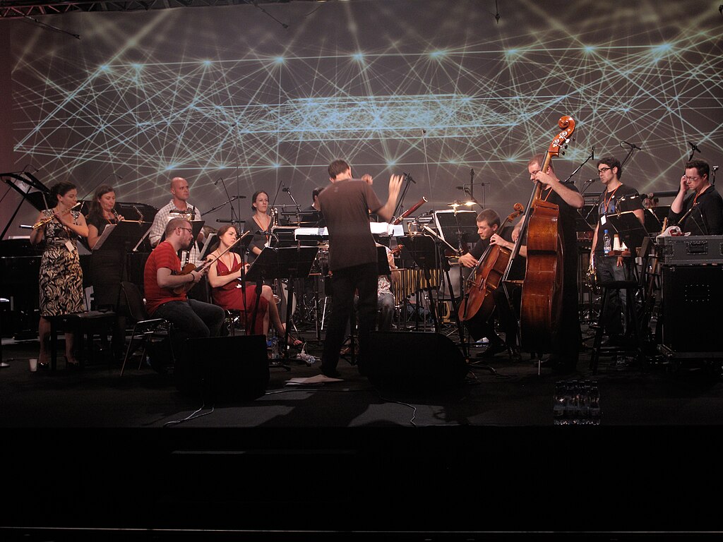 A group of musicians with various instruments performs on stage with a conductor in front of an abstract projection of lines.