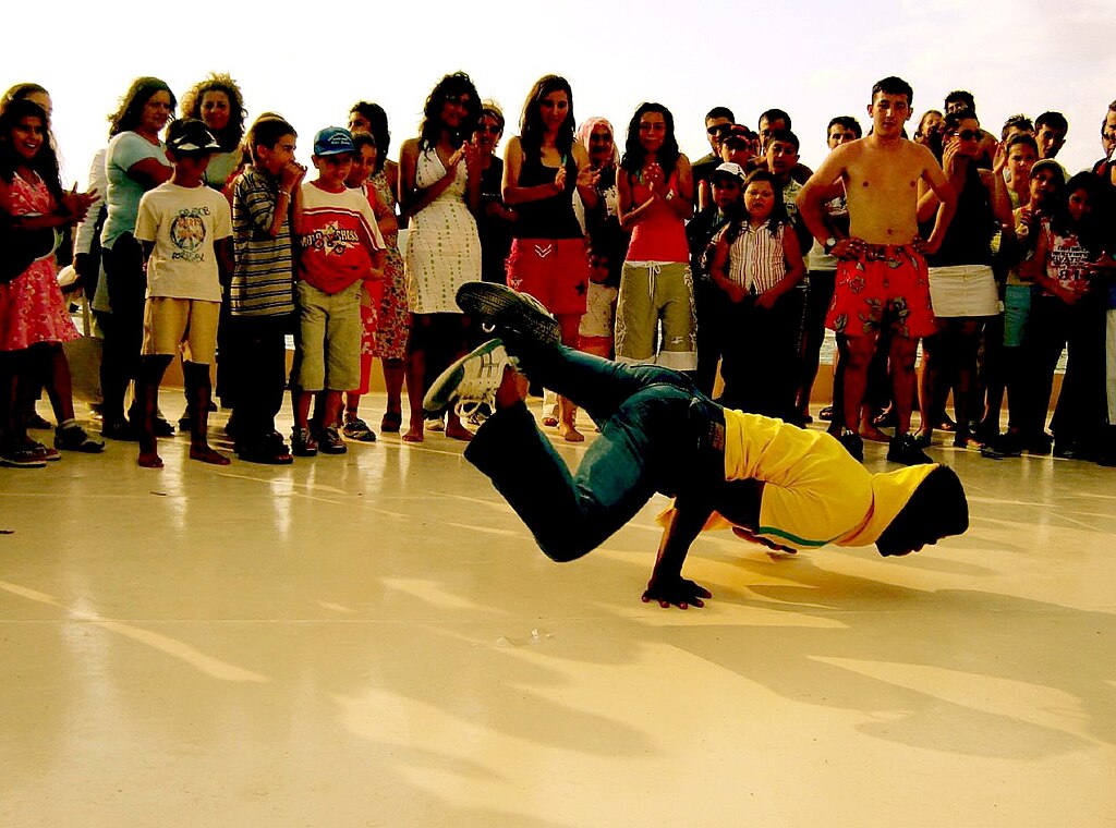 A breakdancer in a yellow shirt performs on the ground surrounded by a crowd of onlookers.