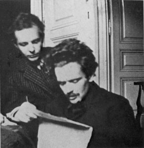 Two men indoors, one seated reading a paper, the other standing behind.