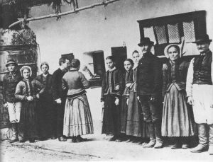 A group of eleven people stands outside a rustic building, with two operating a phonograph on a ledge.