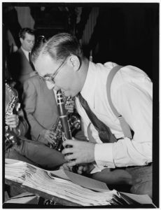 A man with glasses playing a clarinet, accompanied by other musicians in the background.