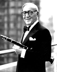 An older man in a tuxedo holding a clarinet, with a blurred cityscape in the background.
