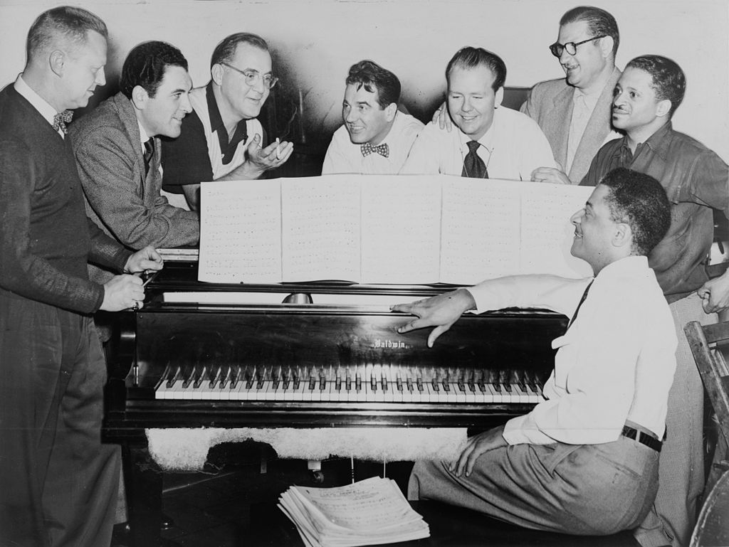 Eight men gather around a grand piano, with sheet music open on the stand.