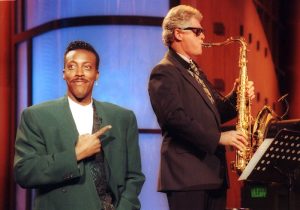 Two people on stage, one pointing and smiling, the other playing a saxophone.