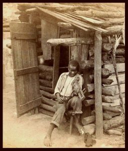 A boy sits on a stool outside a log cabin, holding a homemade stringed instrument.