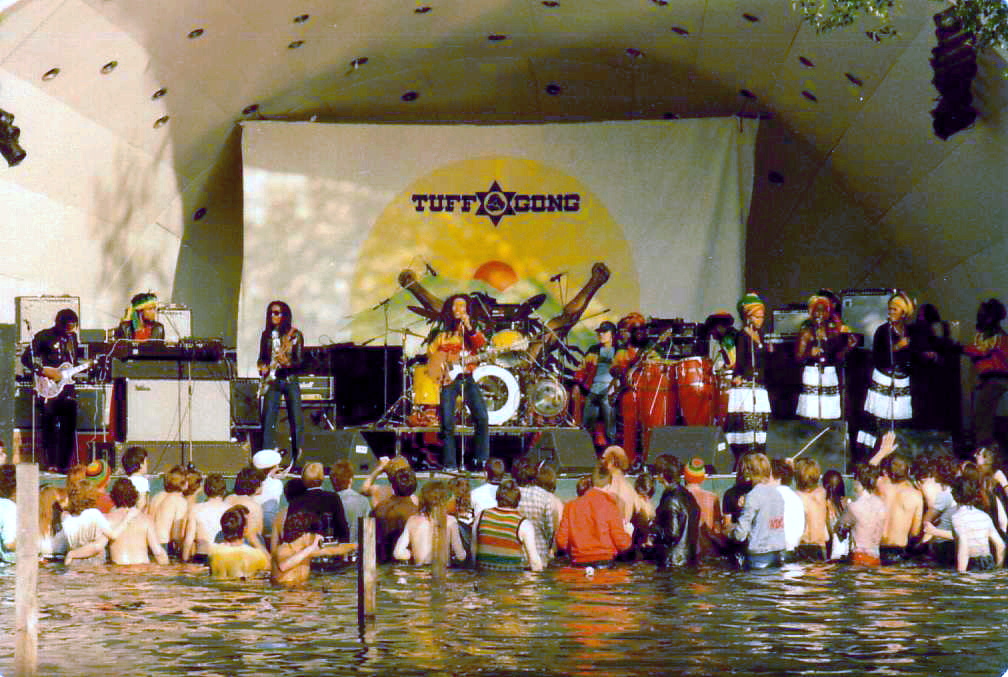 Outdoor concert with musicians performing on a stage above water, watched by a half-submerged audience. Backdrop says "Tuff Gong."