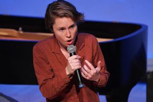 Person speaking into a microphone in front of a grand piano.