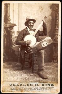 A sepia-toned photograph of a smiling person sitting on a chair playing a banjo, with an ornate fireplace in the background.