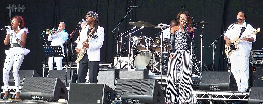 Live performance on stage with six musicians in varied outfits, including two vocalists and multiple instrumentalists.