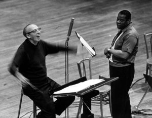 Two men in a music rehearsal, one conducting energetically and the other standing nearby with a focused look.