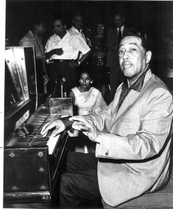 Man playing a grand piano at a social gathering with people in the background.