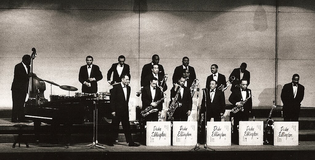 A jazz band performs on stage in tuxedos, featuring saxophonists with "Duke Ellington" music stands, a pianist, and a double bass player.