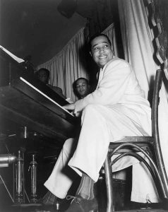 Man in a light suit playing piano with two others in the background.