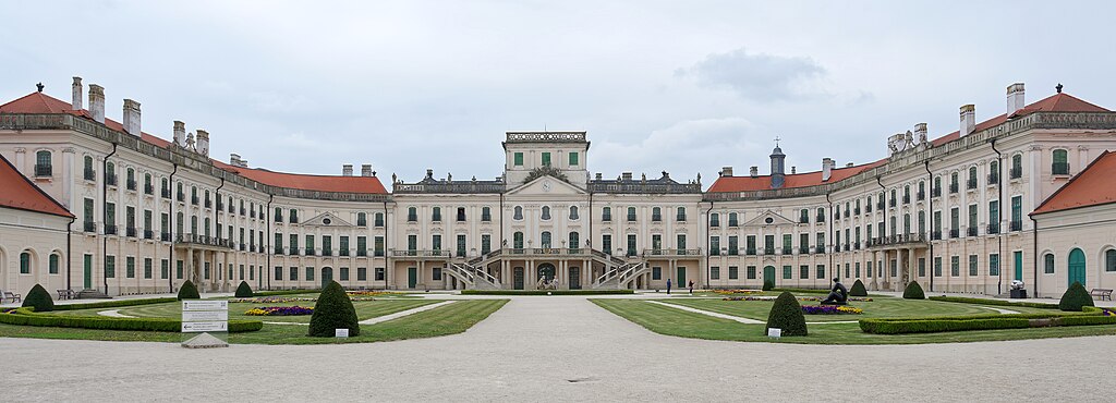 A neoclassical palace with symmetrical wings and a central courtyard, featuring manicured lawns and flower beds.