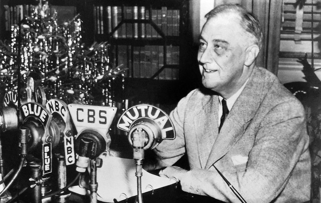 A man in a suit speaks into microphones labeled with various radio station names.