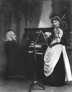 Two musicians performing, one playing the piano and the other playing the violin, in a richly decorated room.