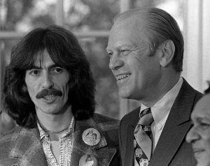 Two men standing and engaging in conversation; one is in casual attire with badges, the other in a suit and tie. A third man's face is barely visible in the lower right corner of the image.